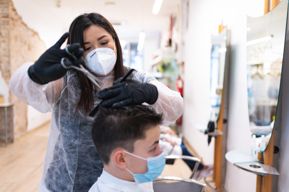 Woman wearing personal protective equipment and a face mask combing and cutting the hair of a boy wearing a face mask