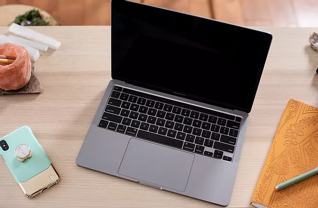 A teal and gold cellphone with a PopSocket, a grey laptop, and a burnt yellow journal sitting on a desk