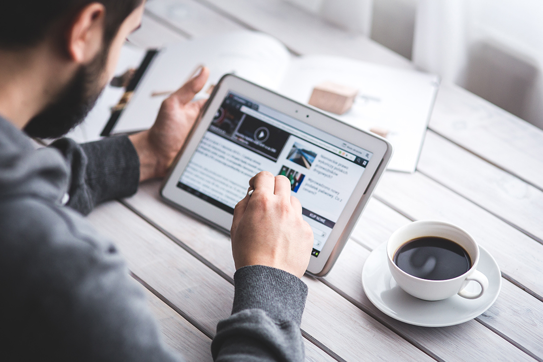 Bearded man sitting at a table using a tablet with a cup of coffee next to him.