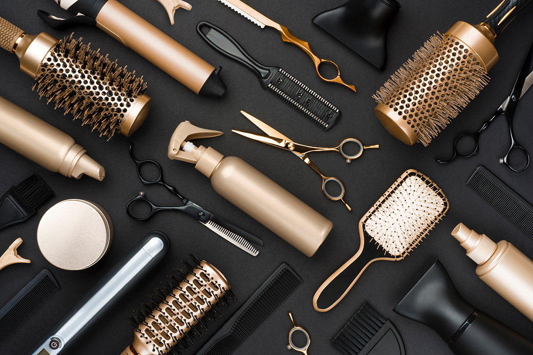 Variety of gold-colored salon tools laid out on a black table—hairbrushes, hair-cutting shears, spray bottles, and more.