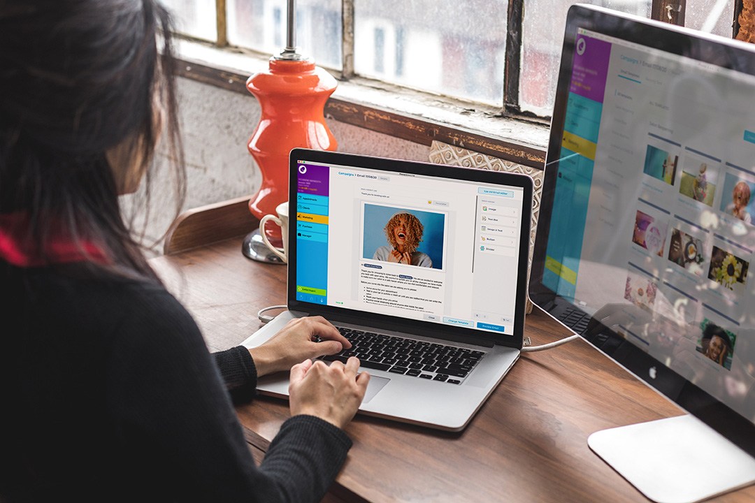 Over the shoulder shot of a woman working on a laptop and desktop monitor using the Phorest Salon Software.