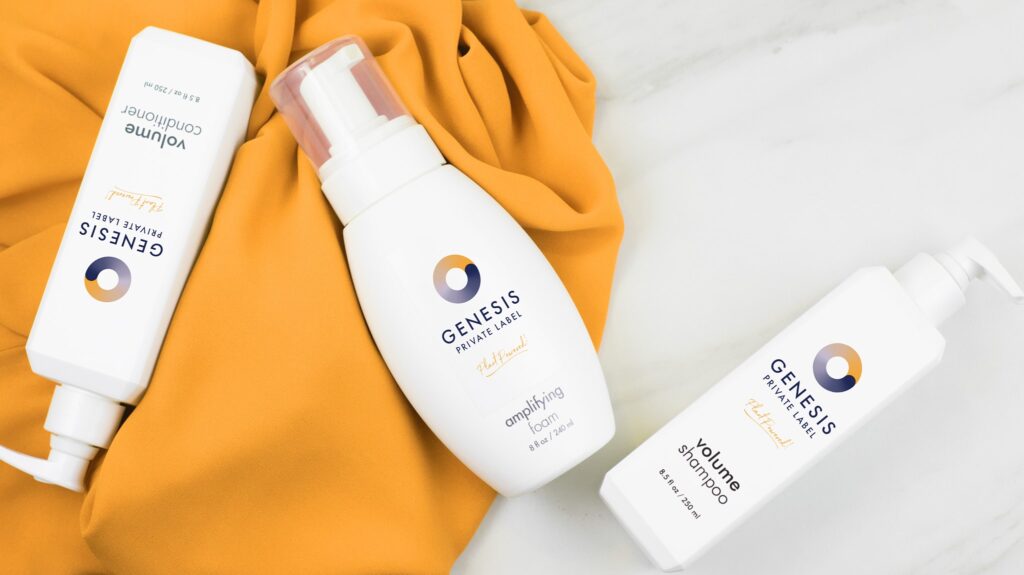 Three white bottles of Genesis Private Label hair products laying on a burnt yellow fabric and white countertop